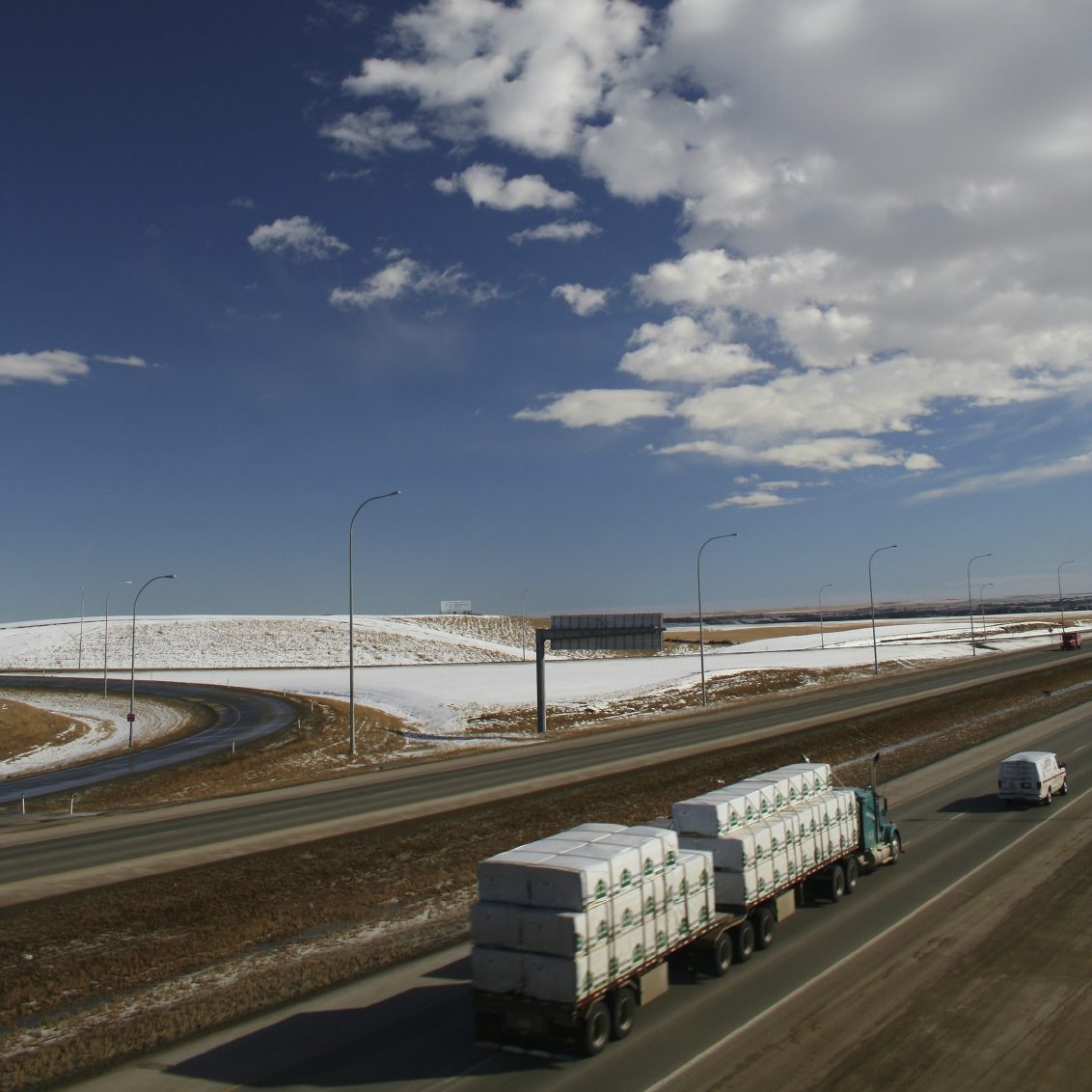 Landschaft mit schneebedeckten Hügeln und Lkw auf einer Straße unter einem blauen Himmel.